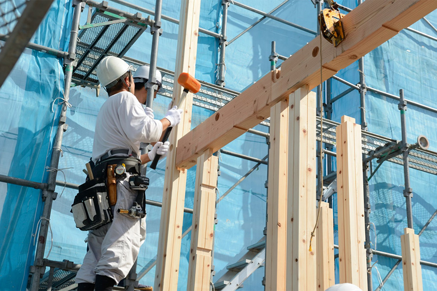 青空の下、建て方の骨組みの上で、職人が木槌を振っている